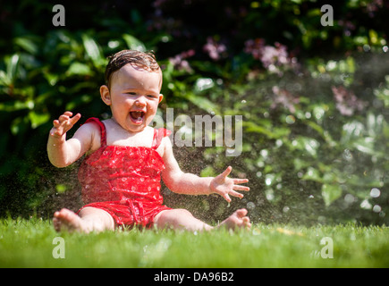 Un bébé crie de plaisir alors qu'elle se refroidit avec de l'eau pulvérisée à partir d'un tuyau flexible. Banque D'Images