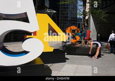 Un travailleur de la ville fume une cigarette comme une femme s'étire les muscles ischio-jambiers après l'exercice suivant pour une installation artistique intitulée "Un à zéro (les dix chiffres)" par l'artiste pop américain Robert Indiana (b 1928), dans la région de Lime Street, City de Londres, la capitale le Square Mile, et son coeur financier. Situé dans le Square Mile, son coeur financier, sont des bureaux environnants et le siège social de la finance et de l'assurance, notamment d'être à proximité du bâtiment de la Lloyds de Londres. Banque D'Images