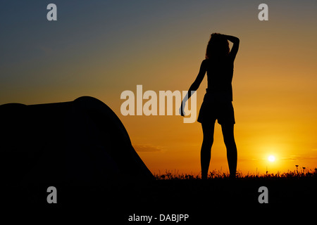 Silhouette d'une femme debout près d'une tente de camping tout en regardant le coucher du soleil. Banque D'Images