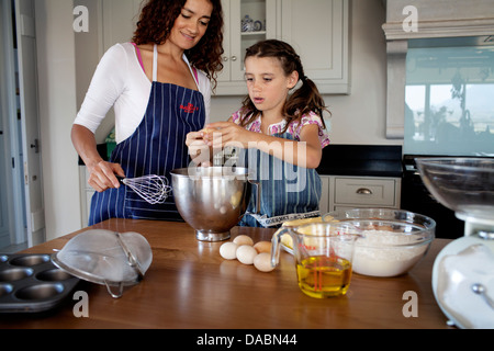 Jeune fille se casse un oeuf dans un mélange à gâteau dans un bol comme mère regarde sur dans la cuisine entouré par des ingrédients - Stonehurst estate Cape Banque D'Images