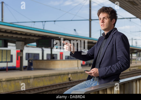 Jeune homme dans une gare en pointant à l'écart et la tenue d'un smartphone Banque D'Images