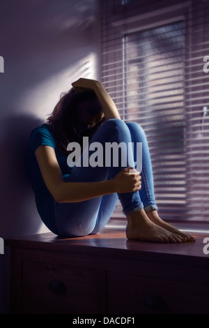 Une adolescente, main sur sa tête, assis par une fenêtre avec la lumière. Banque D'Images