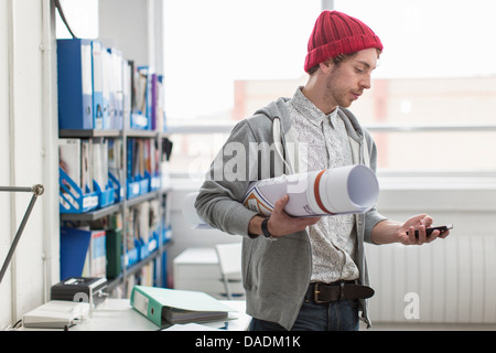 Young office worker holding plans et de l'utilisation de mobile phone in creative office Banque D'Images