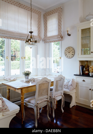 Chaises en rotin peint en blanc et simple table en pin en face de baies vitrées avec stores pâle en salle blanche Banque D'Images