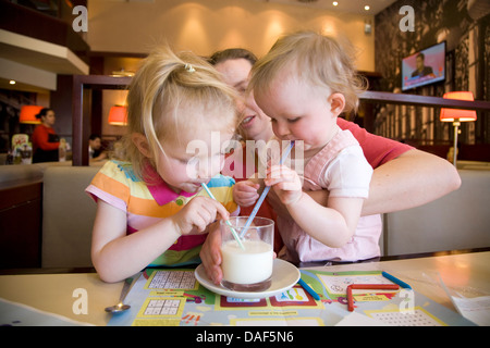 Deux 2 enfants - enfant et bébé - verre de lait de vache avec paille / pailles, tenu par la mère dans un café / restaurant. Banque D'Images