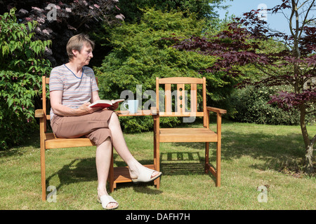 Hauts femme veuve de baby-boomers à la triste et solitaire assis seul à côté d'un compagnon vide siège dans un jardin ensoleillé en été. UK Banque D'Images