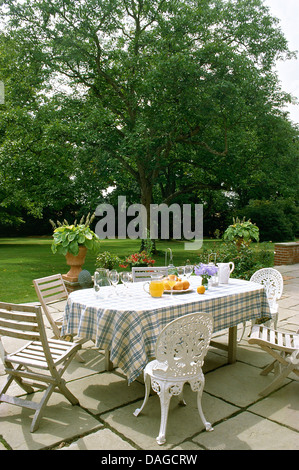 Sol en bois et de chaises en fer forgé blanc à table avec un chiffon bleu vérifié sur un patio dans le jardin de pays Banque D'Images