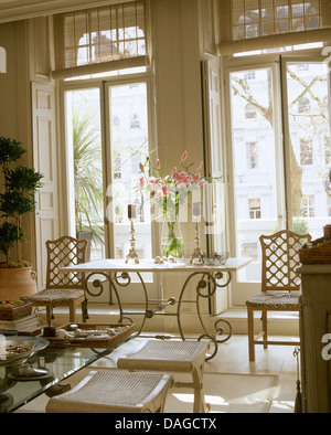 Vase de fleurs rose sur le côté en fer forgé table en face de grand balcon fenêtres avec volets crème en ville Chambre salon Banque D'Images