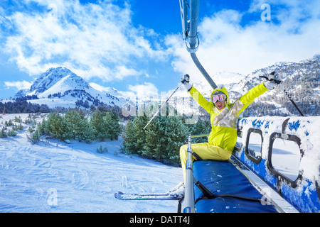 Happy young woman sitting on le remonte-pentes chaise avec les mains levées et sourire avec des montagnes en arrière-plan Banque D'Images