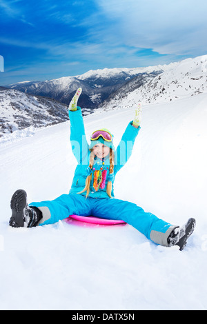 Happy girl faisant glisser en descente sur traîneau avec ses mains levées, le port de masque de ski, avec les montagnes en arrière-plan Banque D'Images