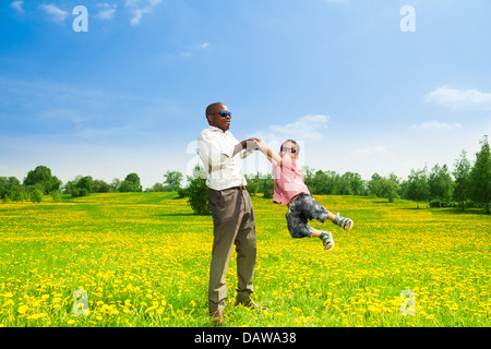 Heureux père noir avec son fils faisant tourner son garçon sur le terrain avec pissenlits jaunes dans le parc Banque D'Images