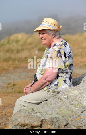 Une vieille dame portant un chapeau de paille assis sur des roches à Guernesey Banque D'Images