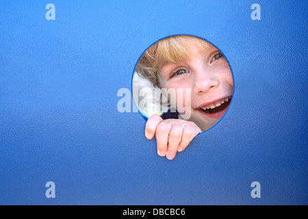 Un cute blonde enfant est sourire et rire comme il jette sa tête à travers un trou dans un tunnel de jeu un jour d'été Banque D'Images