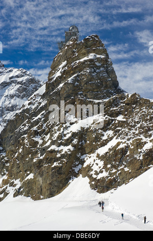 Les touristes à pied à travers le Mönchsjoch Hut de navigation au haut de la partie supérieure du Glacier d'Aletsch, avec l'Observatoire du Sphinx topping le 3571 mètres (11 716 pieds) Sphinx peak, Jungfraujoch, Alpes Bernoises, près de Grindelwald, Suisse Banque D'Images