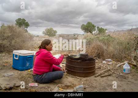 Une femme sans-abri sur un maïs cuisiniers grill extérieur primitif primitive un campement de plein air de la ville désertique de Victorville, CA. Banque D'Images