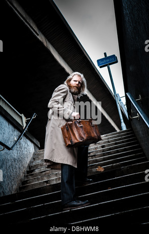 Homme barbu en costume et manteau tenant un vieux cartable en cuir à monter pas hors d'un passage inférieur et à la hâte. Banque D'Images