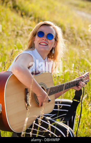 Teenage girl wearing lunettes noires à jouer de la guitare dans les champs. À l'extérieur. L'été. Banque D'Images