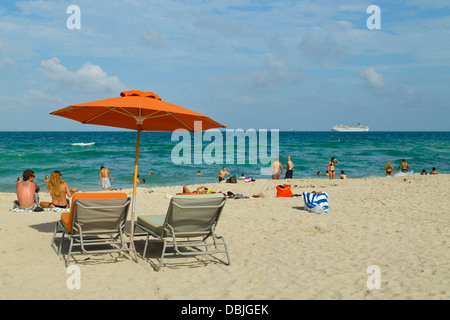 Chaises de plage, parasol orange et beaucoup d'heureux amateurs de plage à South Beach, Miami, Floride. Un bateau de croisière est dans la distance. Banque D'Images