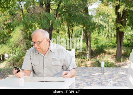 Personnes âgées man texting on his mobile assis à l'extérieur, dans l'ombre d'un arbre essayant de lire l'écran avec ses lunettes de lecture Banque D'Images