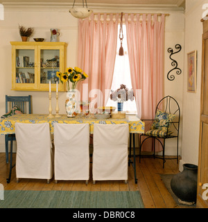 Meuble blanc-couvre sur des chaises à table avec un chiffon jaune dans la salle à manger de style économie avec rideaux rose Banque D'Images