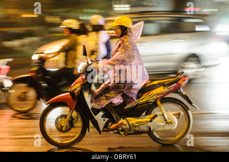 Moto Honda Wave Casque Jaune Poncho de Pluie Flou de Mouvement Vieille Quartier Hanoi Vietnam | HANOI, Vietnam — Une femme portant un poncho translucide navigue dans la circulation rendue glissante par la pluie dans le Vieille Quartier sur une Honda Wave. Introduite au début des années 2000 pour concurrencer les importations bon marché, la série Wave est rapidement devenue le véhicule de prédilection des navetteurs au Vietnam. Son châssis étroit et son moteur à quatre temps fiable restent particulièrement adaptés pour manœuvrer dans le réseau de rues dense et labyrinthique de la capitale. Banque D'Images