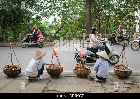 Vendeurs de rue vendre des ignames Hanoi Vietnam // HANOI, Vietnam — deux vendeurs de rue à Hanoi, Vietnam, vendent des ignames et d'autres racines sur le bord d'une route comme les gens sur des scooters courent. Banque D'Images