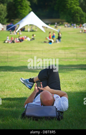 / Man lying on grass en utilisant un téléphone mobile à l'avant-plan. Dans la distance et tente ouverte. Journée ensoleillée Banque D'Images