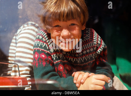 Jeune garçon souriant à la caméra regardant par une fenêtre dans les années 1970 Norfolk Royaume-Uni Angleterre Grande-Bretagne dans les années 1970 1973 KATHY DEWITT Banque D'Images