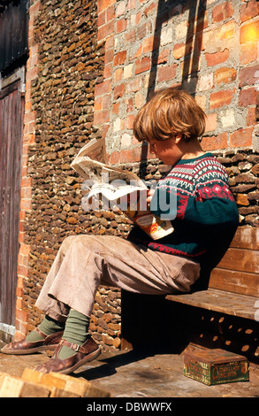 Jeune garçon britannique aux cheveux de gingembre portant des sandales et un pull Fair isle assis sur un banc lisant un magazine de bandes dessinées dans les années 1960 ou 1970 Norwich, Norfolk Angleterre Royaume-Uni Grande-Bretagne KATHY DEWITT Banque D'Images