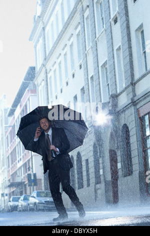 Businessman talking on cell phone under umbrella in rainy street Banque D'Images