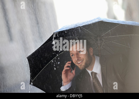 Businessman talking on cell phone under umbrella in rain Banque D'Images