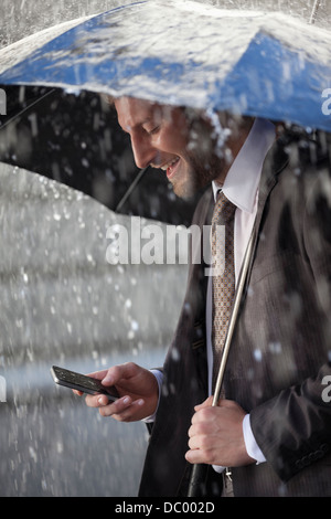 Businessman text messaging on cell phone under umbrella in rain Banque D'Images