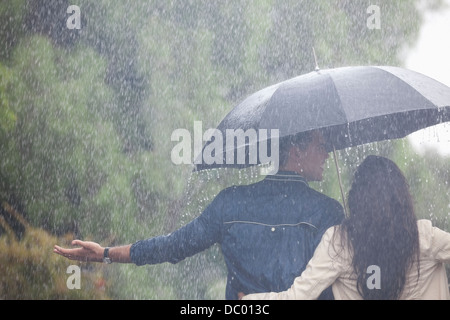Couple walking with arms outstretched under umbrella in rain Banque D'Images