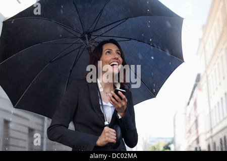 Happy businesswoman text messaging with cell phone under umbrella in rain Banque D'Images