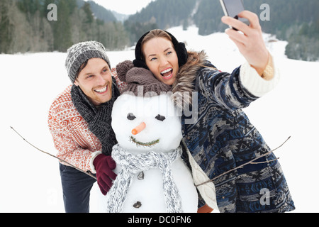 Enthousiaste couple taking self-portrait with snowman in snowy field Banque D'Images