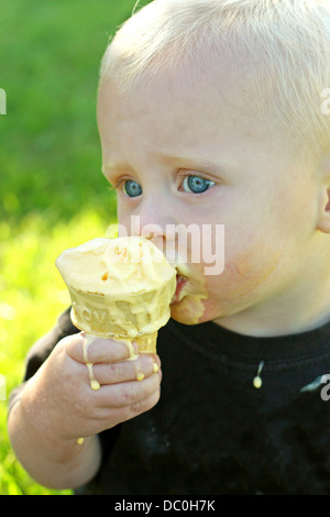 Un mignon, blond bébé garçon est assis dehors dans l'herbe un jour d'été, une alimentation désordonnée, de fusion de la glace à la vanille cone Banque D'Images