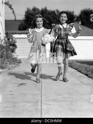 Années 1930 Années 1940 DEUX FILLES SAUTANT VERS LE BAS TROTTOIR HOLDING HANDS LOOKING AT CAMERA Banque D'Images