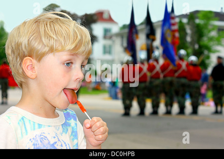 Un enfant est à un défilé. léchant un meunier de bonbons, comme les militaires tenant des drapeaux américains en mars Banque D'Images