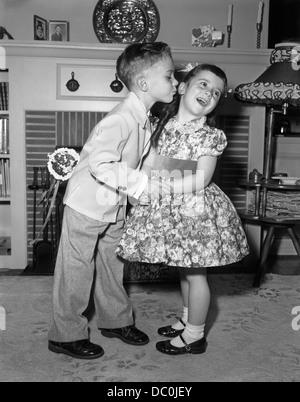 J1950s petit garçon en costume et cravate essayant d'embrasser petite fille en robe de soirée Banque D'Images