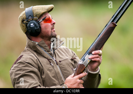 L'homme à tirer sur une simulation de tir jeu de tir d'argile portant chapeau de tweed, de l'oreille et des lunettes de protection Banque D'Images