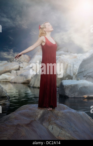 Une femme en robe rouge est debout sur un rocher dans l'eau et profiter du soleil