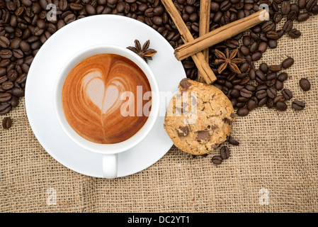 Tasse de café avec de la mousse en forme de coeur avec des grains de café et les bâtons de cannelle et d'anis étoilé et de biscuit sur fond de toile de jute Banque D'Images