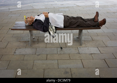 Gardant son bâton de marche, un vieux monsieur dort sur un banc de la rue de ville dans le centre de Londres, ses lunettes à charnière en position haute. Banque D'Images