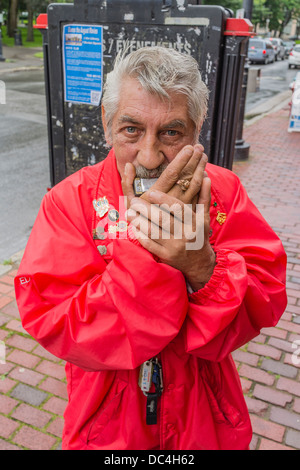 Un vieux mendiant adultes joue de l'harmonica mâles dehors sur le trottoir dans une veste rouge vif, et les cheveux gris. Banque D'Images