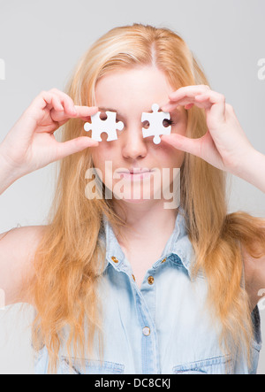 Jeune femme blonde tenant deux pièces de puzzle blanc devant ses yeux Banque D'Images