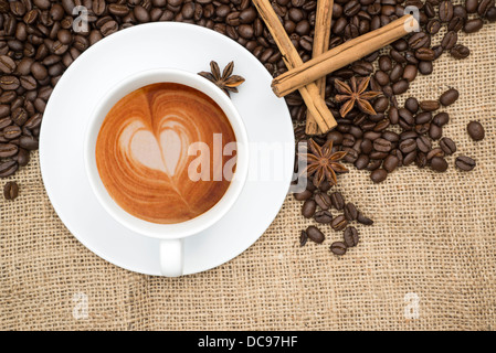 Tasse de café avec de la mousse en forme de coeur avec des grains de café et les bâtons de cannelle et d'anis étoilé sur fond de toile de jute Banque D'Images