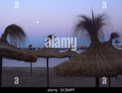 Des parasols de paille et la pleine lune dans la baie de Cala Estancia, Majorque, Espagne. Banque D'Images