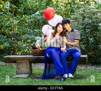 Photo de young adult couple sitting on log banc, à l'avant, avec des verres remplis de vin rouge qui se tient dans les mains Banque D'Images