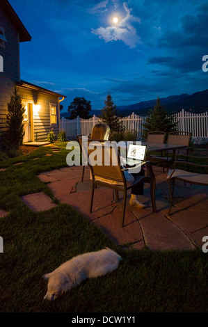 Femme travaillant à l'extérieur à la maison sur un ordinateur portable au crépuscule, animal chiot Golden Retriever sur la pelouse Banque D'Images