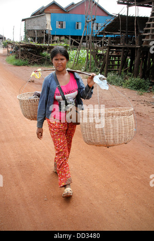 Dame cambodgienne portant des paniers sur chemin de terre dans la région de Kampong Khleang devant des maisons sur pilotis au Cambodge Banque D'Images